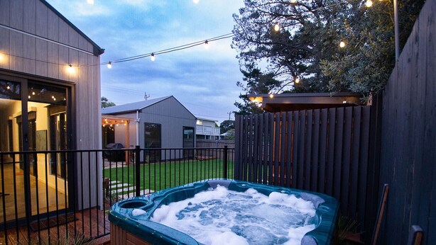 A hot tub in a backyard with a wooden fence, string lights, and a house in the background.