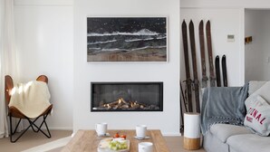 A living room with a fireplace, a painting of waves, skis on the wall, and a cozy seating area. - Avalon: a striking five-bedroom, three-bathroom holiday rental. (Jindabyne)