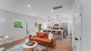 A modern living room with a sofa, coffee table, and dining area. A kitchen with a dining table and chairs is visible in the background. - New Stylish Townhome in Cotswold with Ensuite Baths, Chef's Kitchen & EV Charger (Charlotte)