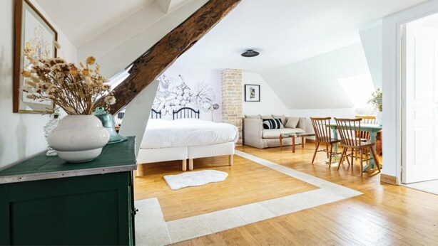 A bedroom with a sloped ceiling, a bed, a dresser, a vase with dried flowers, a dining area with chairs and a table, and a view of the outdoors.