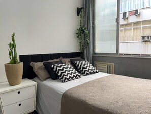 Een netjes opgemaakt bed met patroon kussens, een potplant en een nachtkastje met lades.