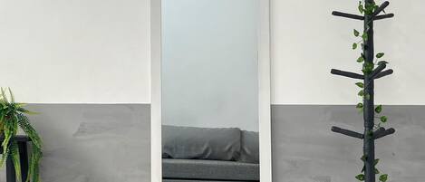 A mirror with a gray sofa in the reflection, a black plant stand with green foliage, and a gray wall.