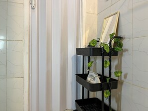 Un baño con pared de azulejos blancos, un estante negro con un espejo y plantas verdes, y un toallero blanco.