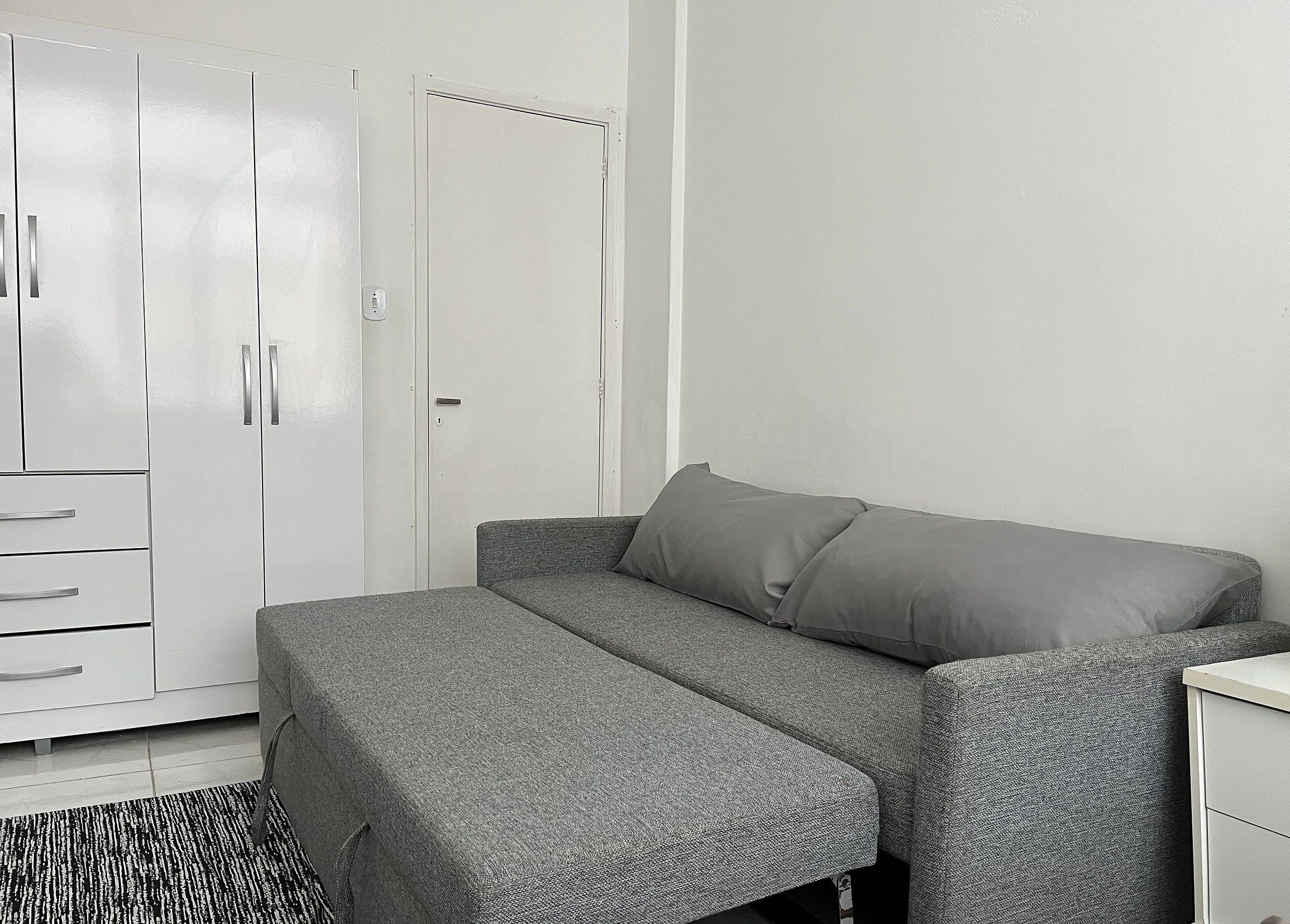 A compact room with a grey sofa bed, a white wardrobe, and a small bedside table.