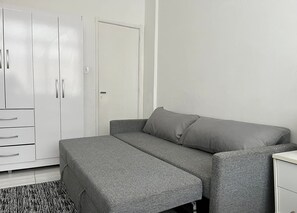 Een compacte kamer met een grijze slaapbank, een witte kledingkast en een klein nachtkastje.