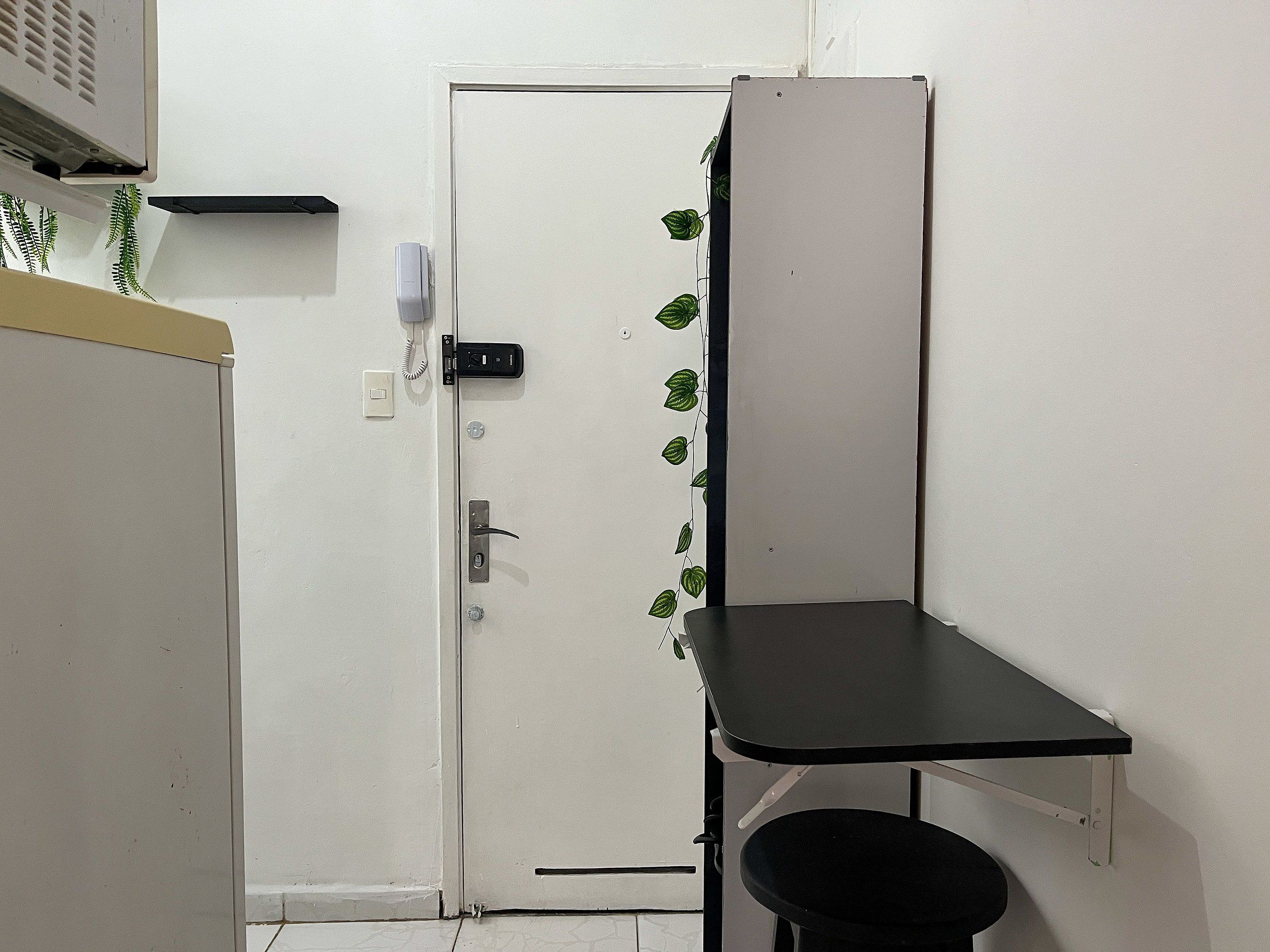 Une pièce avec une porte blanche, un petit bureau avec une chaise noire et une plante accrochée au mur.