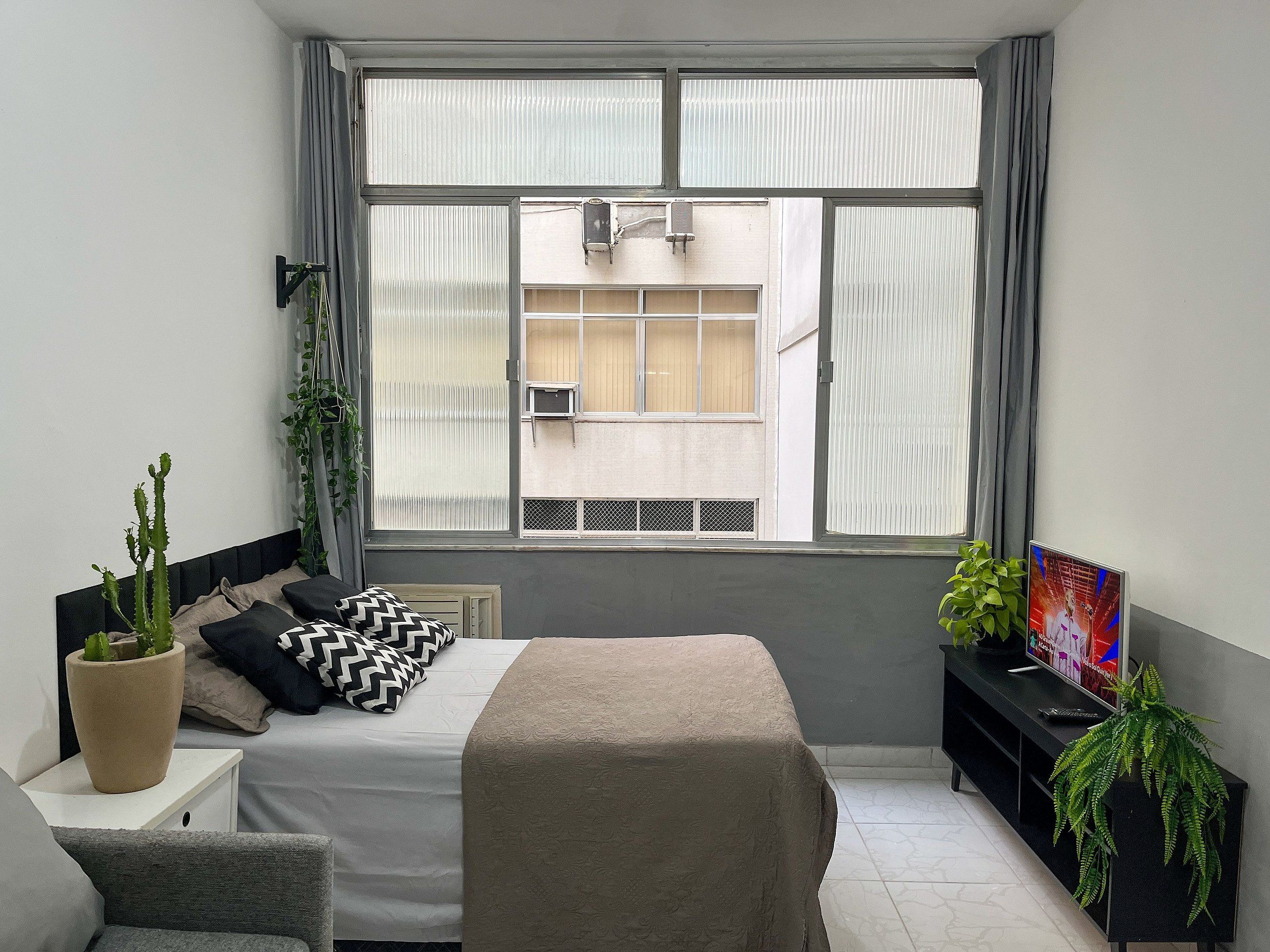 Un dormitorio con cama, ventana con cortinas, televisor y planta en maceta.