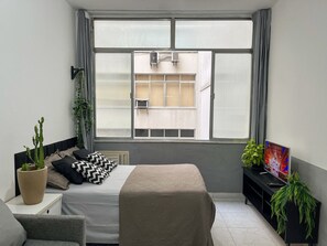 Un dormitorio con cama, ventana con cortinas, televisor y planta en maceta.