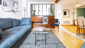 Un salon moderne avec un canapé gris, une table basse en verre, un tapis à motifs, une bibliothèque en bois et une grande fenêtre avec des rideaux.