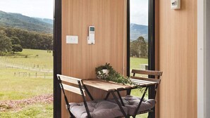 A room with a wooden bench and cushioned chairs, a small table with a plant, and a view of a green landscape with hills. - Paddock & Peak by Tiny Away (Wesburn)