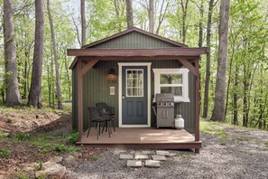 A small cabin with a door, a window, a grill, and two chairs outside. - Tiny Cabin w Fire Pit Game Room and Dog Friendly (Great Cacapon)