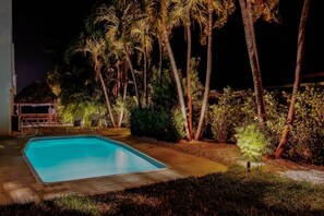 Una piscina rodeada de palmeras y una estructura con techo de paja al caer la noche.