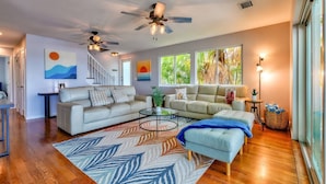 A spacious living room with a large sofa, a smaller sofa, a coffee table, and a patterned rug. There are ceiling fans, a staircase, and large windows with a view of greenery.