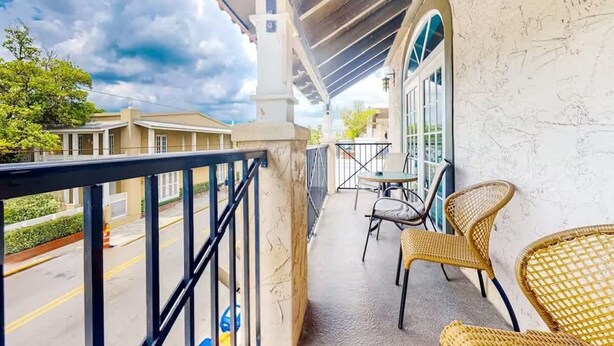 A balcony with wicker chairs, a table, and a view of the street and neighboring buildings. - Coral Reef Suite | 1 bed near Duval & waterfront (Key West)