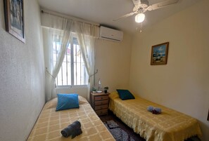 A small, well-lit bedroom with two beds, a window with curtains, a ceiling fan, and a single wall-mounted air conditioner.