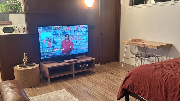 Una sala de estar con televisor, piso de madera, sofá de cuero marrón y una cama roja.