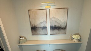 Un pasillo con piso de madera, dos otomanos redondos, una estantería con objetos decorativos y dos cuadros en la pared.
