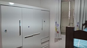 A bedroom with a large wardrobe, a bed with a blue patterned bedspread, and a window with a curtain.