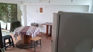 A room with a bed, a table, a stool, a refrigerator, and a potted plant.