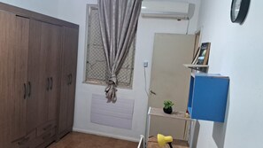 Habitació amb un armari, una finestra amb cortines, un aparell d'aire condicionat, una prestatgeria amb una planta i un rellotge a la paret.