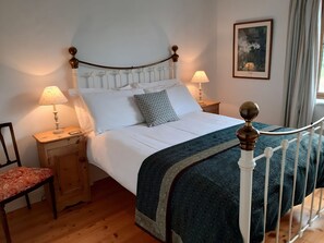 Een netjes opgemaakt bed met witte lakens, een geborduurde deken en een houten hoofdbord. Er staan twee nachtkastjes met lampen, een stoel met een rode kussen, en een ingelijst foto aan de muur.
