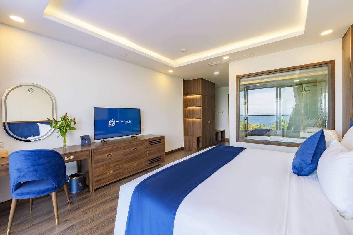 Moderni hotellihuone, jossa on suuri sänky, työpöytä tuolilla, televisio ja suuri ikkuna, josta avautuu näkymä merelle.