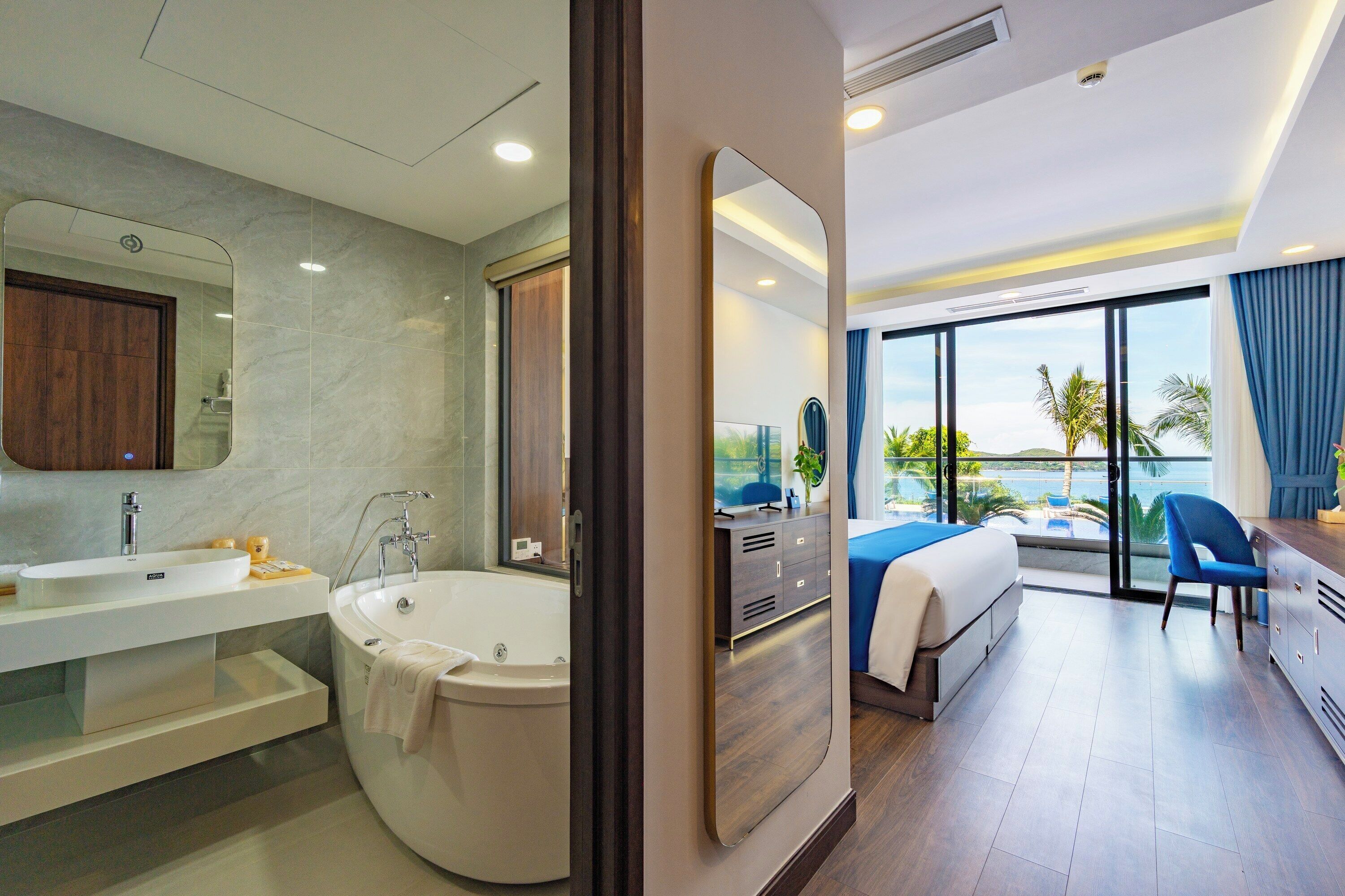 A modern hotel room with a bathroom, a bed, a desk, and a view of the ocean.