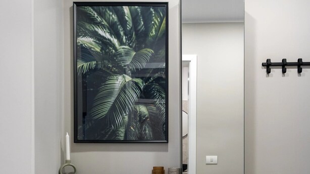A bathroom with a large mirror, a framed tropical leaf artwork, a small round ottoman, and a shelf with toiletries. - Downtown Bucharest Studio Close to Cismigiu & Universitatii (Bucharest)