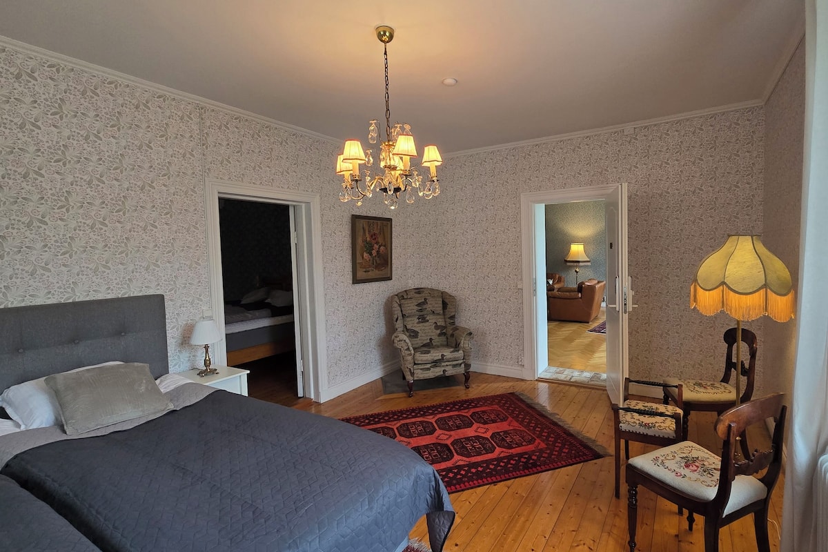A bedroom with a bed, a chair, a nightstand, a lamp, a chandelier, and a door leading to another room.
