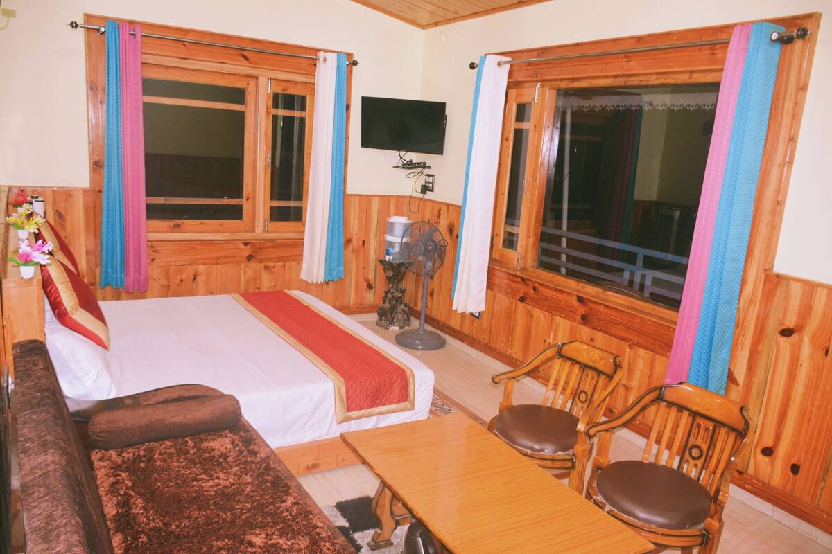Una habitación con cama, una mesa de madera, dos sillas, televisor y ventilador.