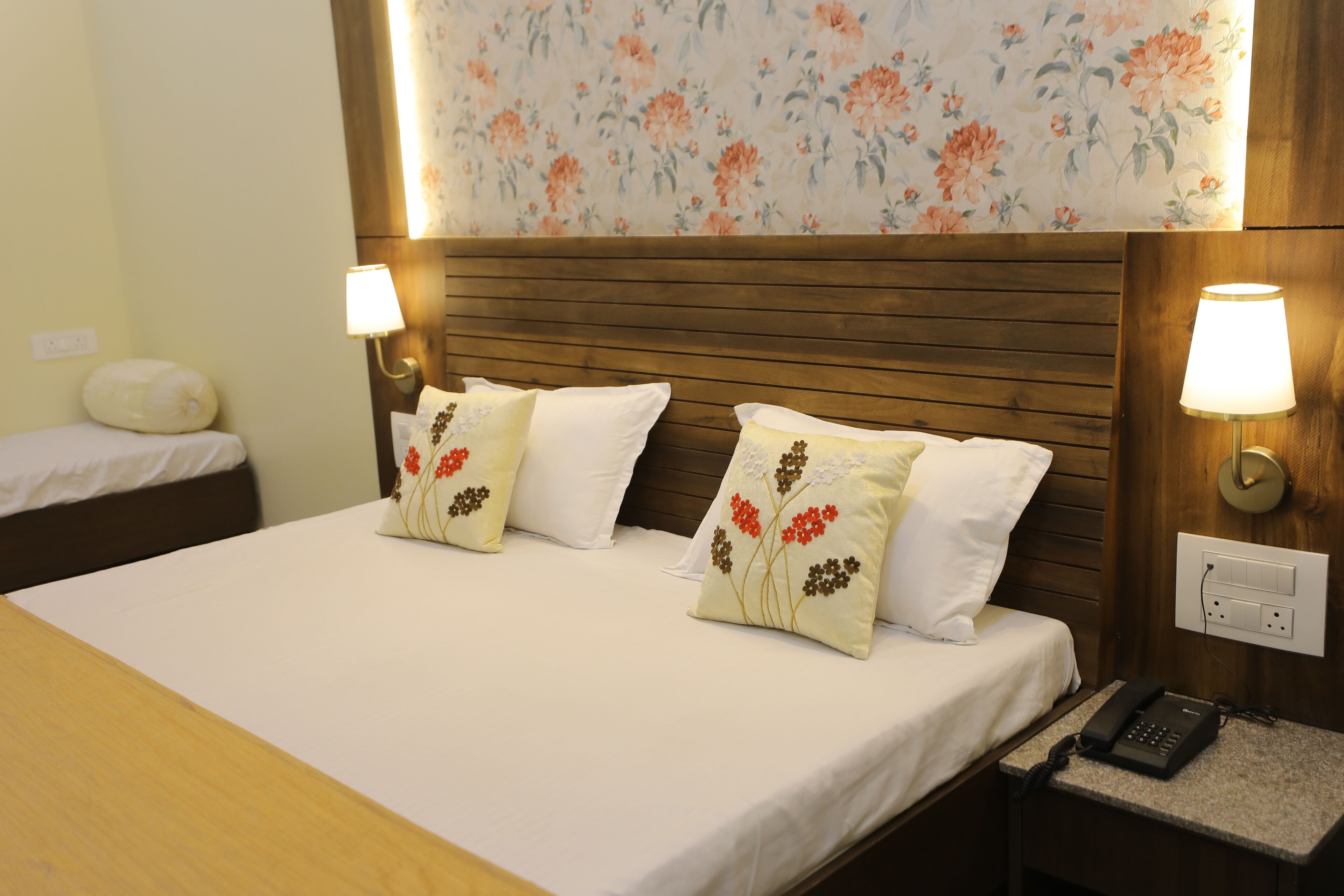 A hotel room with a wooden headboard, two bedside lamps, a telephone, and floral wallpaper.