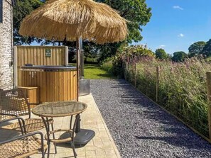 A patio with a thatched umbrella, a wooden barbecue, wicker furniture, and a gravel path.