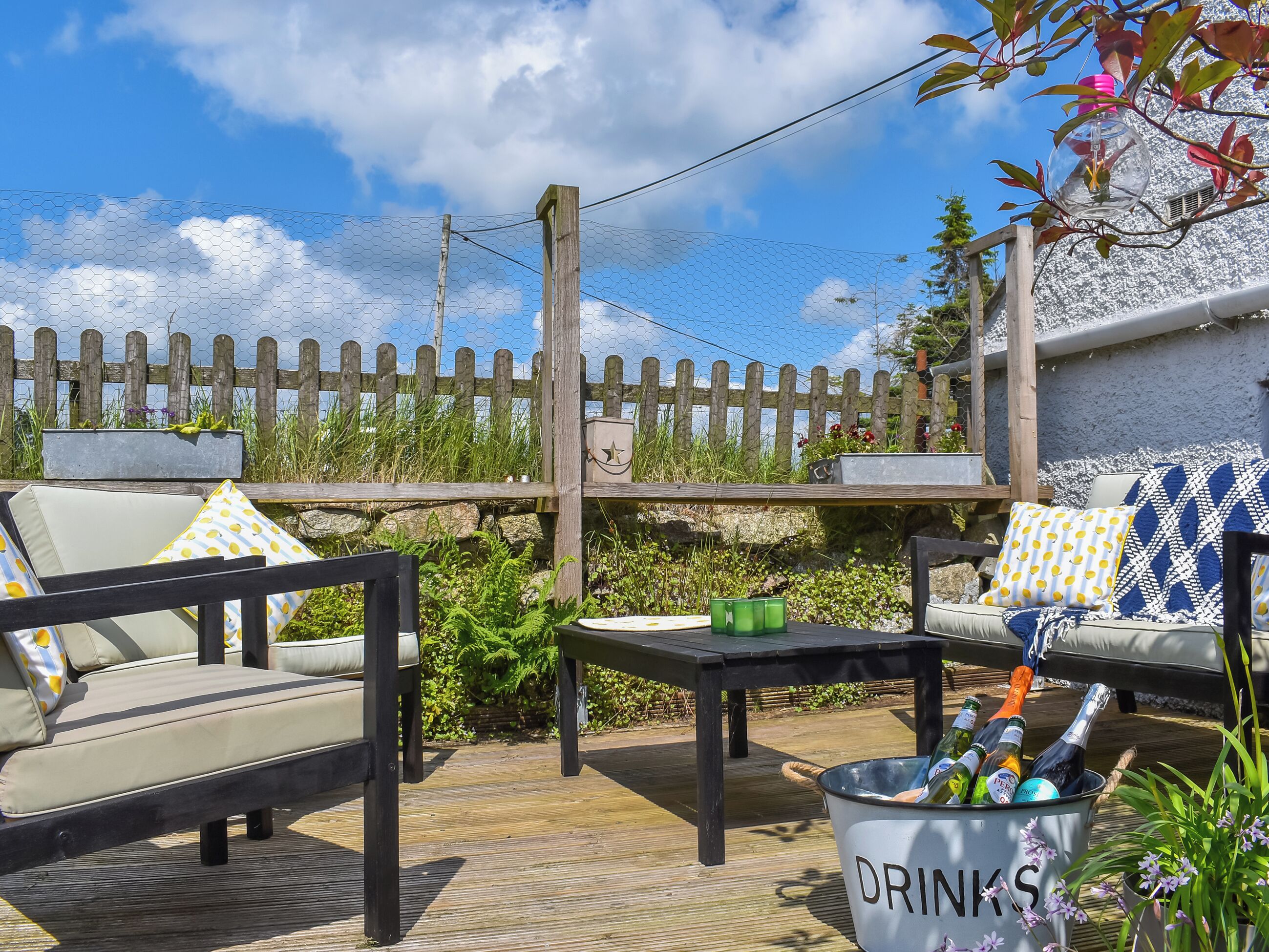 Un espace extérieur aménagé en patio, avec une clôture en bois, une table sur laquelle se trouve un seau à boissons, et deux canapés ornés de coussins à motifs.