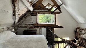 Une pièce avec un plafond en pente, une fenêtre donnant sur la verdure, un lit recouvert d’une couverture blanche et un escalier muni d’une rampe.