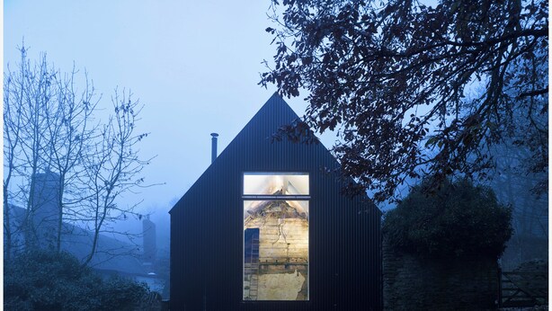 Un bâtiment moderne aux couleurs sombres, doté d’une grande fenêtre et entouré d’arbres et d’un mur de pierre.