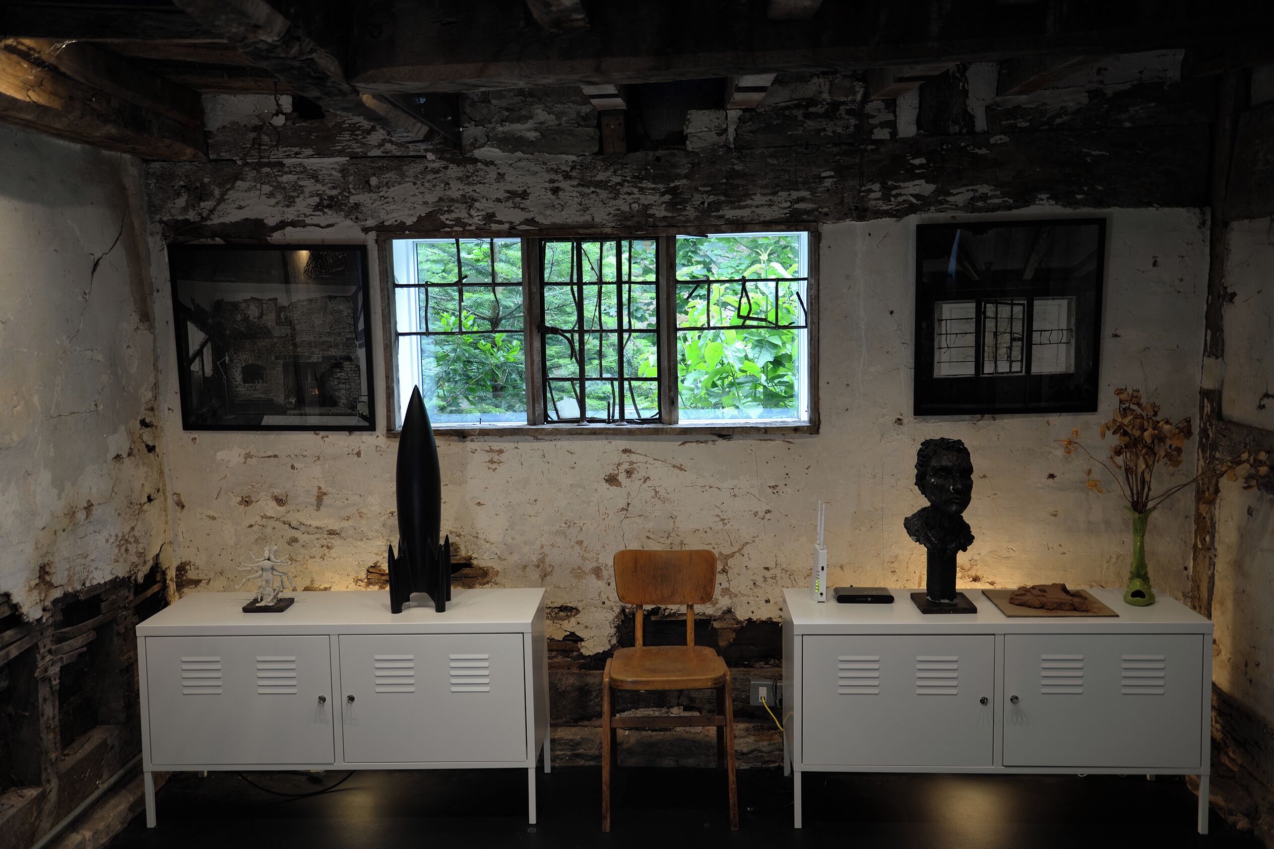 A room with a rustic aesthetic, featuring a wooden chair, a white cabinet with a vase, and a window with a view of greenery.
