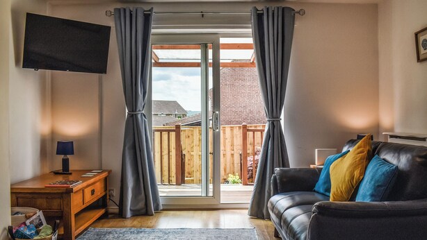 Ein Wohnzimmer mit einer Couch, einem an der Wand befestigten Fernseher, einem Holztisch und Blick durch eine Glastür auf einen Außenbereich.
