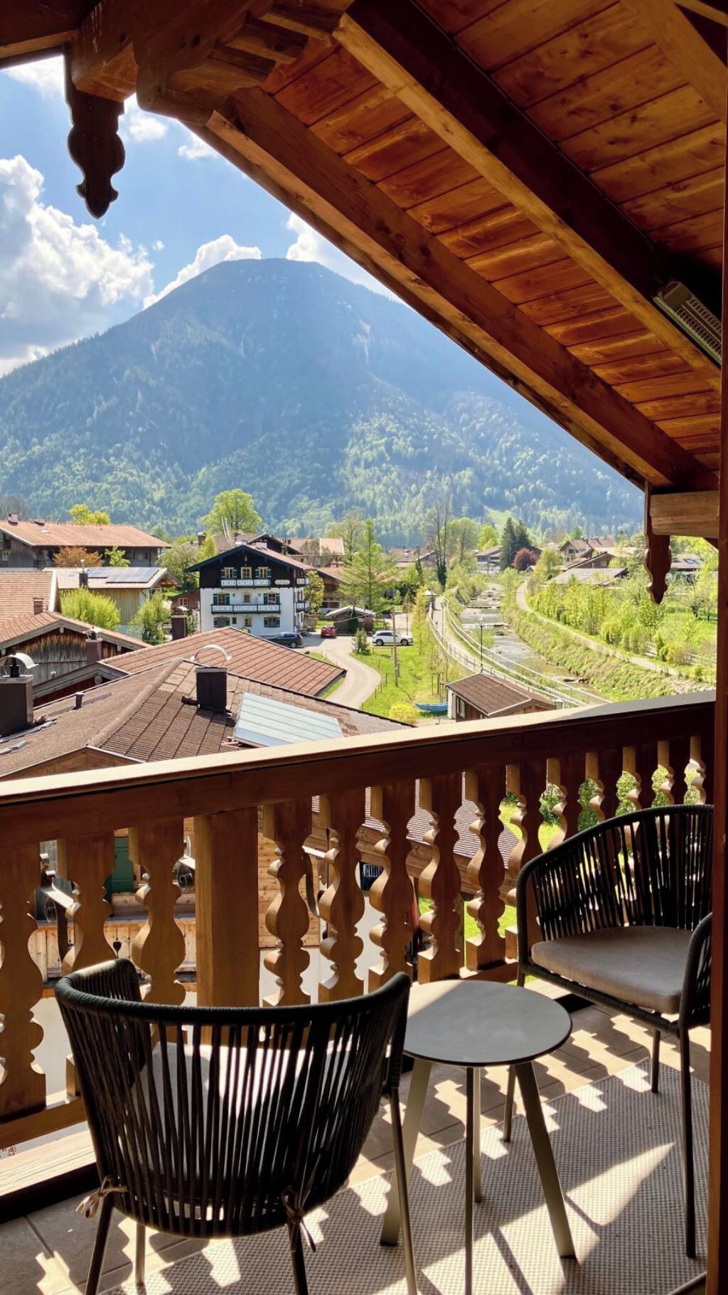 Ein hölzerner Balkon mit zwei Stühlen und einem Tisch, mit Blick auf eine gebirgige Landschaft mit Gebäuden und einer Eisenbahn.