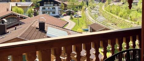 A wooden balcony with two chairs and a table, overlooking a mountainous landscape with buildings and a railway.
