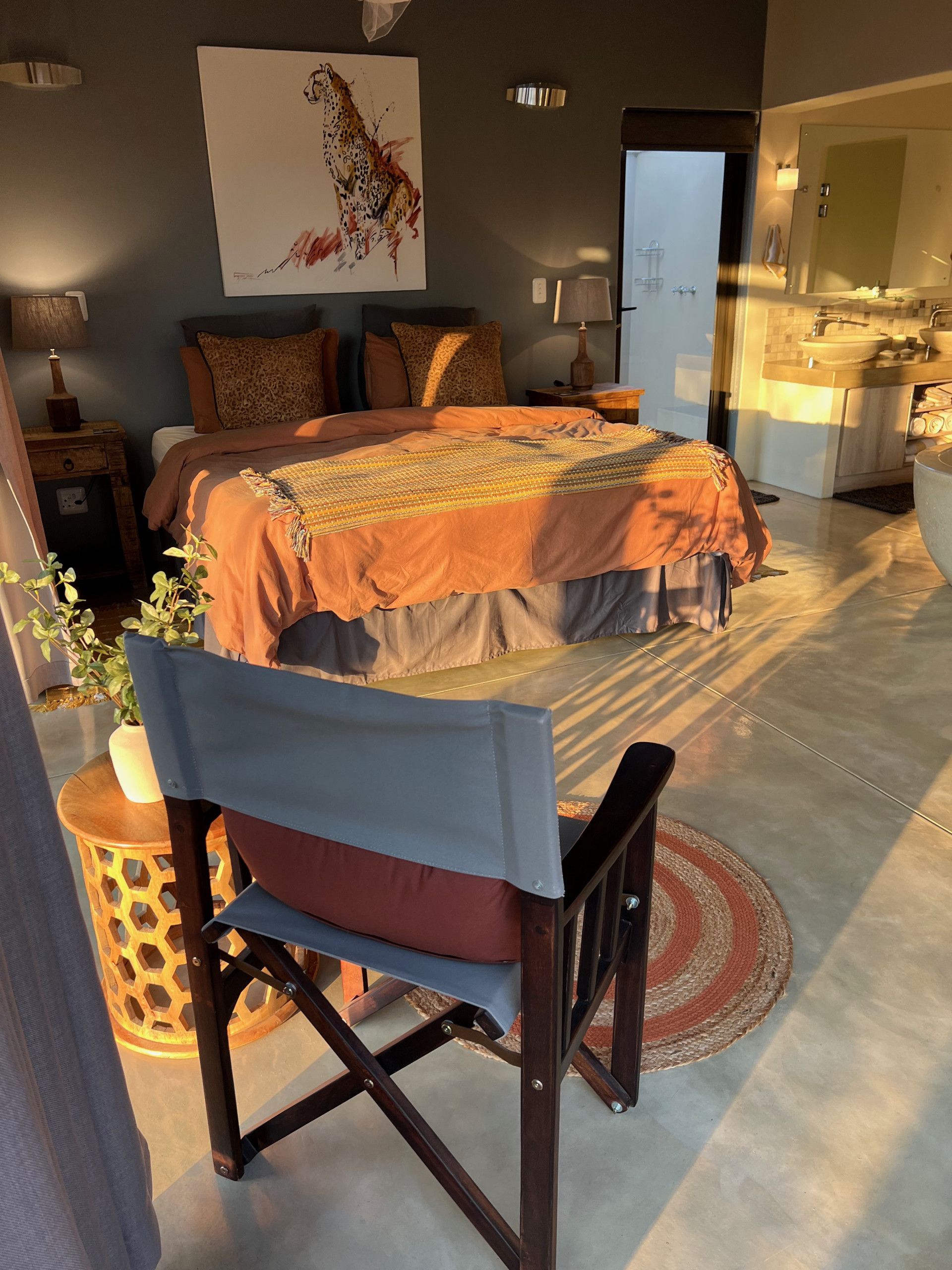 A bedroom with a bed, a nightstand, a lamp, a painting, and a bathroom visible through an open door.