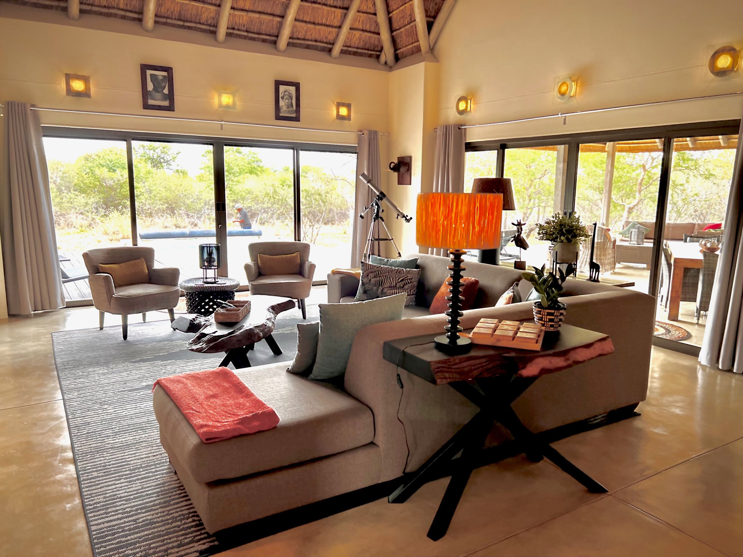 A spacious living area with a sofa, armchairs, a coffee table, and a lamp. Large windows provide a view of the outdoors.