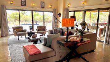 A spacious living area with a sofa, armchairs, a coffee table, and a lamp. Large windows provide a view of the outdoors.