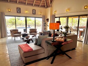 Amplia sala de estar con sofá, sillones, una mesa de centro y una lámpara. Ventanas grandes ofrecen vistas al exterior.