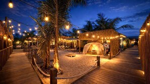 Une terrasse en bois surmontée d’une structure à toit de chaume, agrémentée de guirlandes lumineuses et de palmiers.