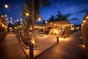 A wooden deck with a thatched-roof structure, string lights, and palm trees.