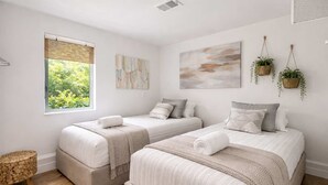 Une chambre avec deux lits, une fenêtre donnant sur un paysage verdoyant, un tabouret en osier et des plantes suspendues.