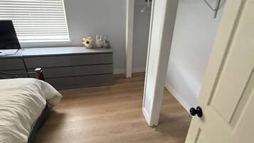 A bedroom with a bed, a desk, a dresser, a window with blinds, and a door leading to another room.