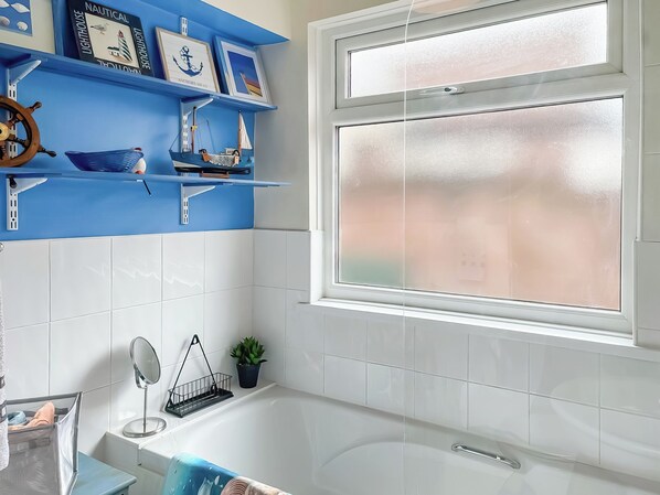 Un baño con bañera, una ventana, un estante decorado con motivos náuticos y una planta en maceta.