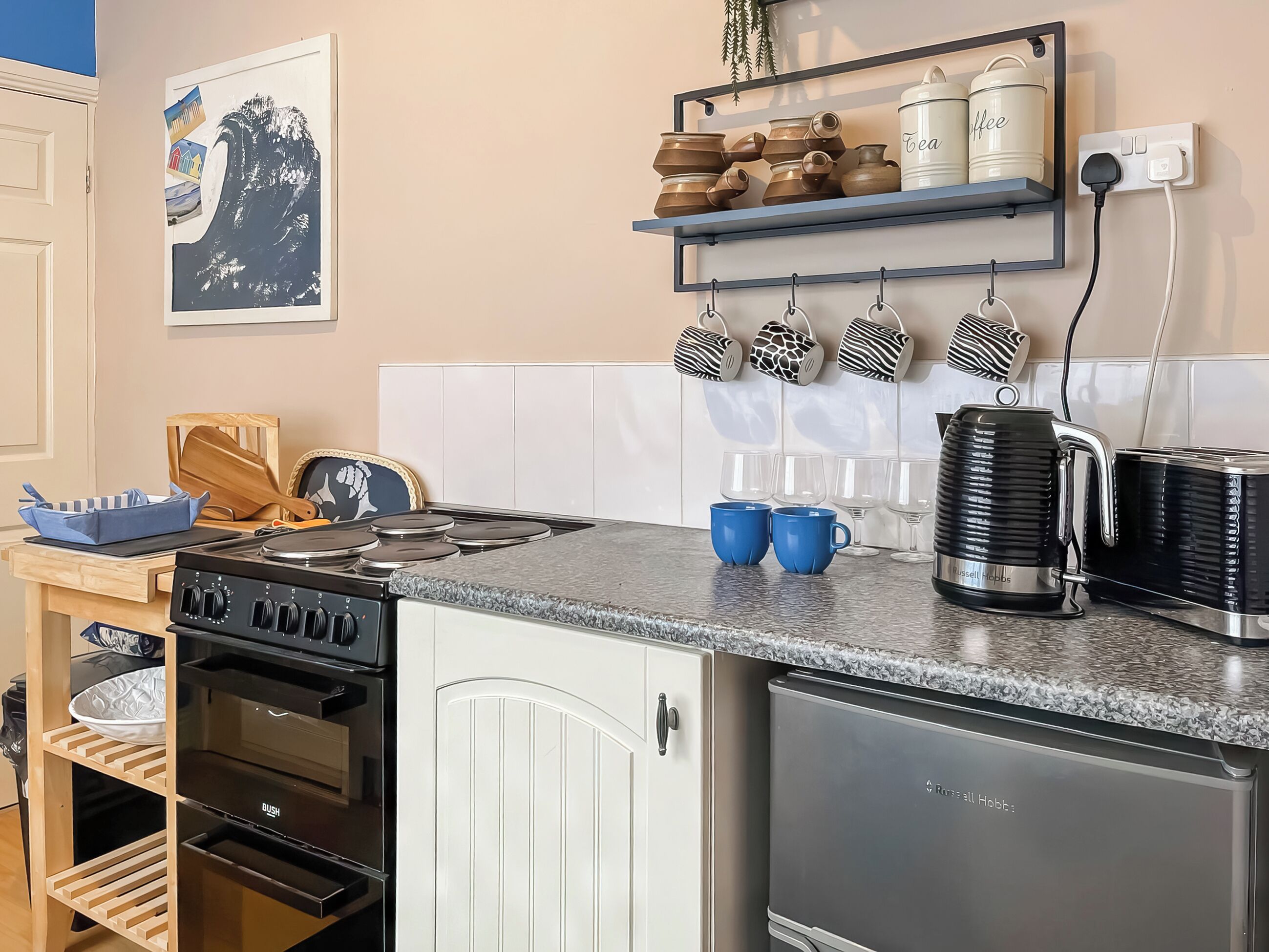 A kitchen with a stove, oven, refrigerator, and a shelf with mugs and tea containers.