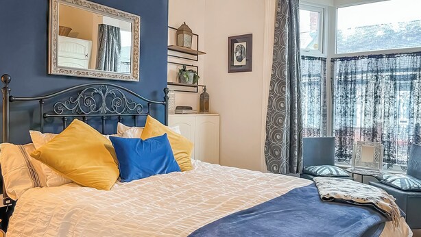 A neatly made bed with a patterned bedspread and pillows, a bedside table with a lamp, a mirror on the wall, and a window with curtains.